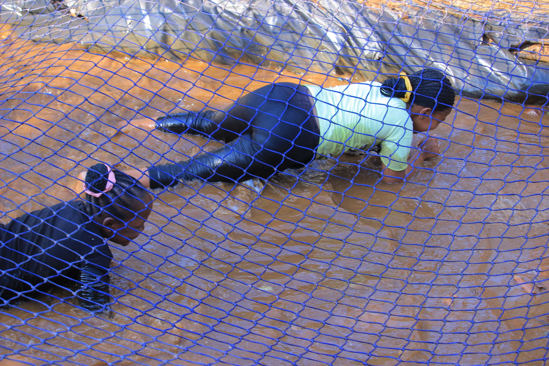 Two learners crawling under a net through muddy water